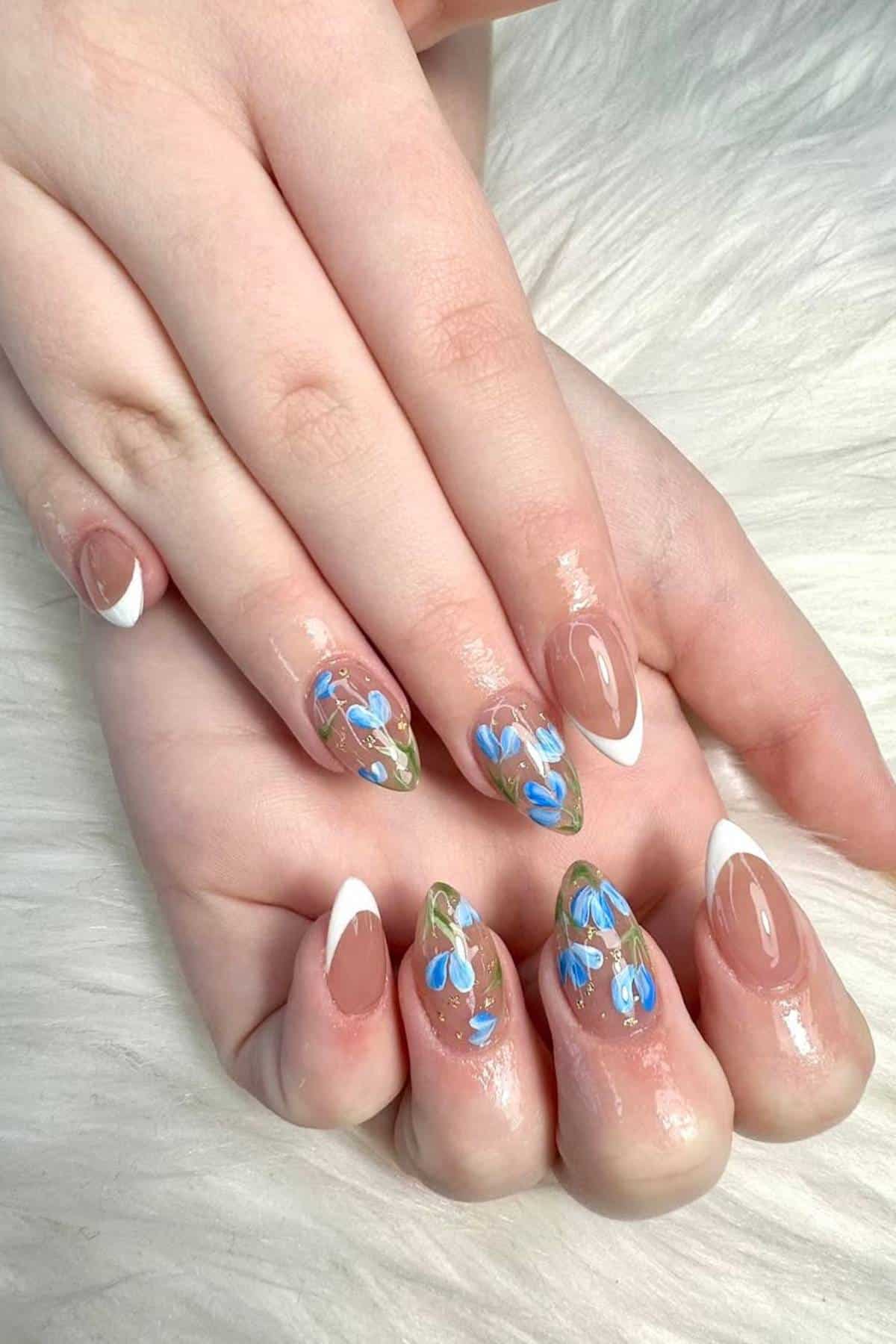 Hands with almond-shaped nails feature a French tip design on some fingers and blue floral nail art with green accents on others, displayed on a white textured surface.