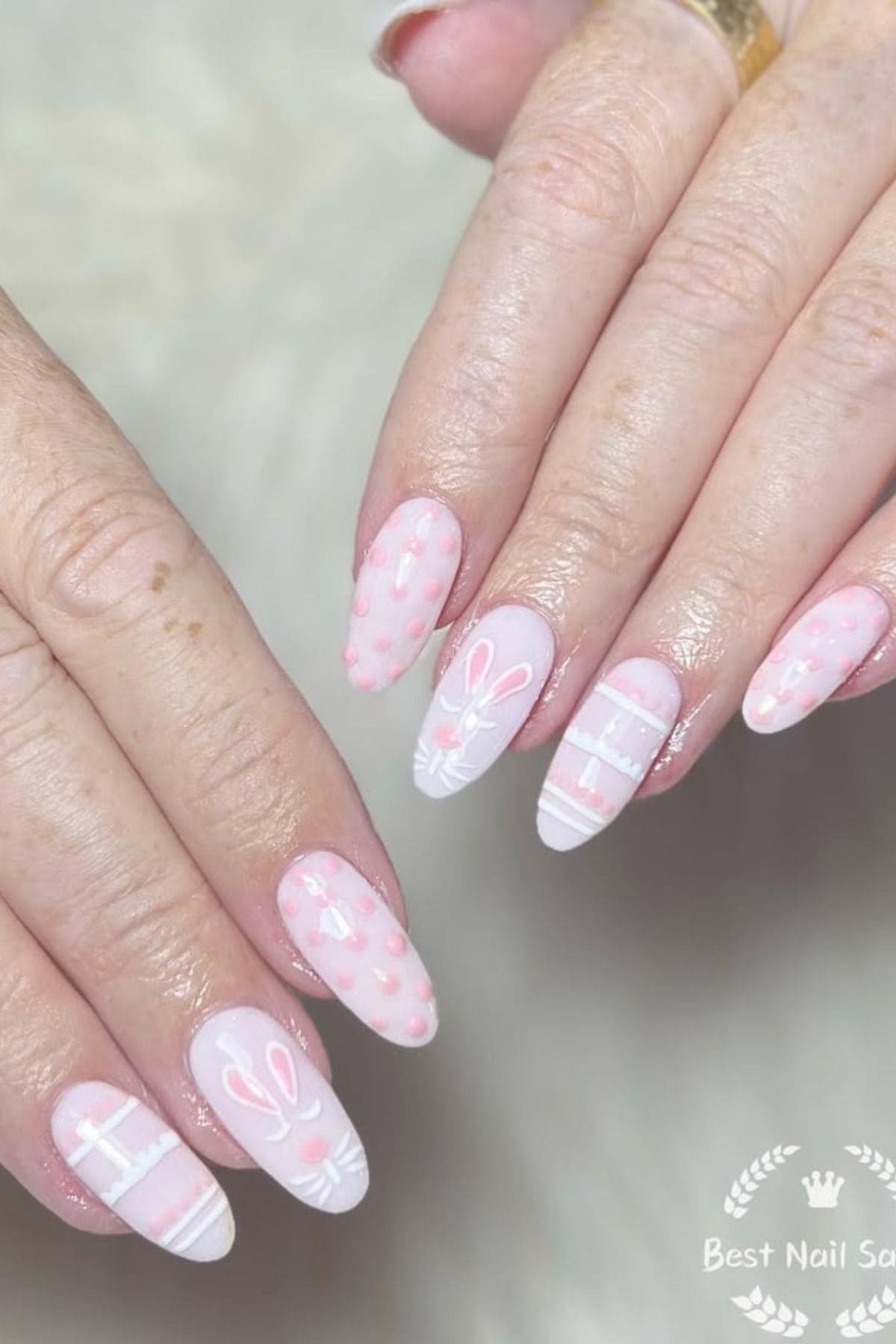 Hands with long, almond-shaped nails painted light pink with white stripes, pink dots, and white bunny face designs on some nails.