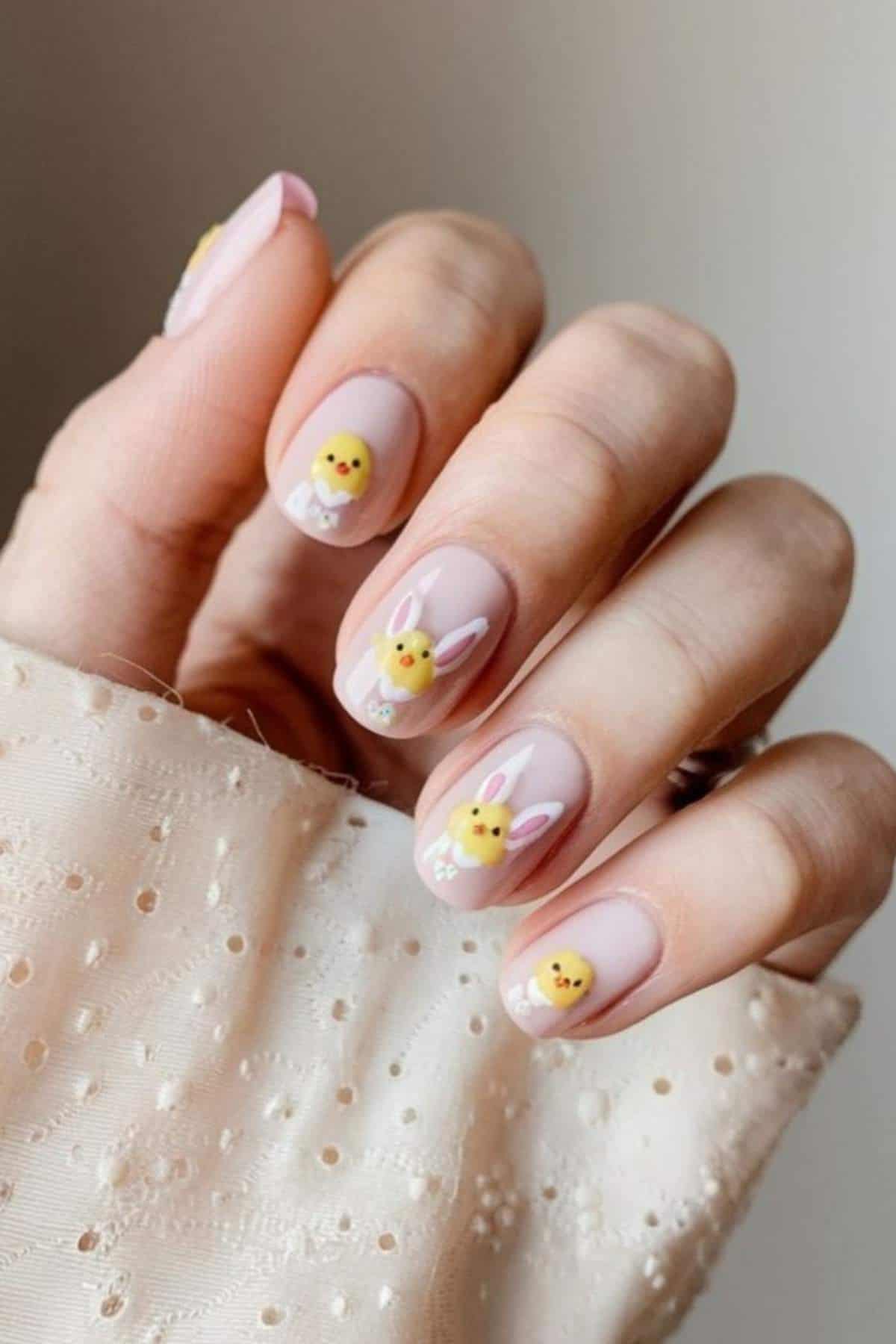 A hand with nude-colored nails featuring small chick and bunny face nail art on each fingernail.