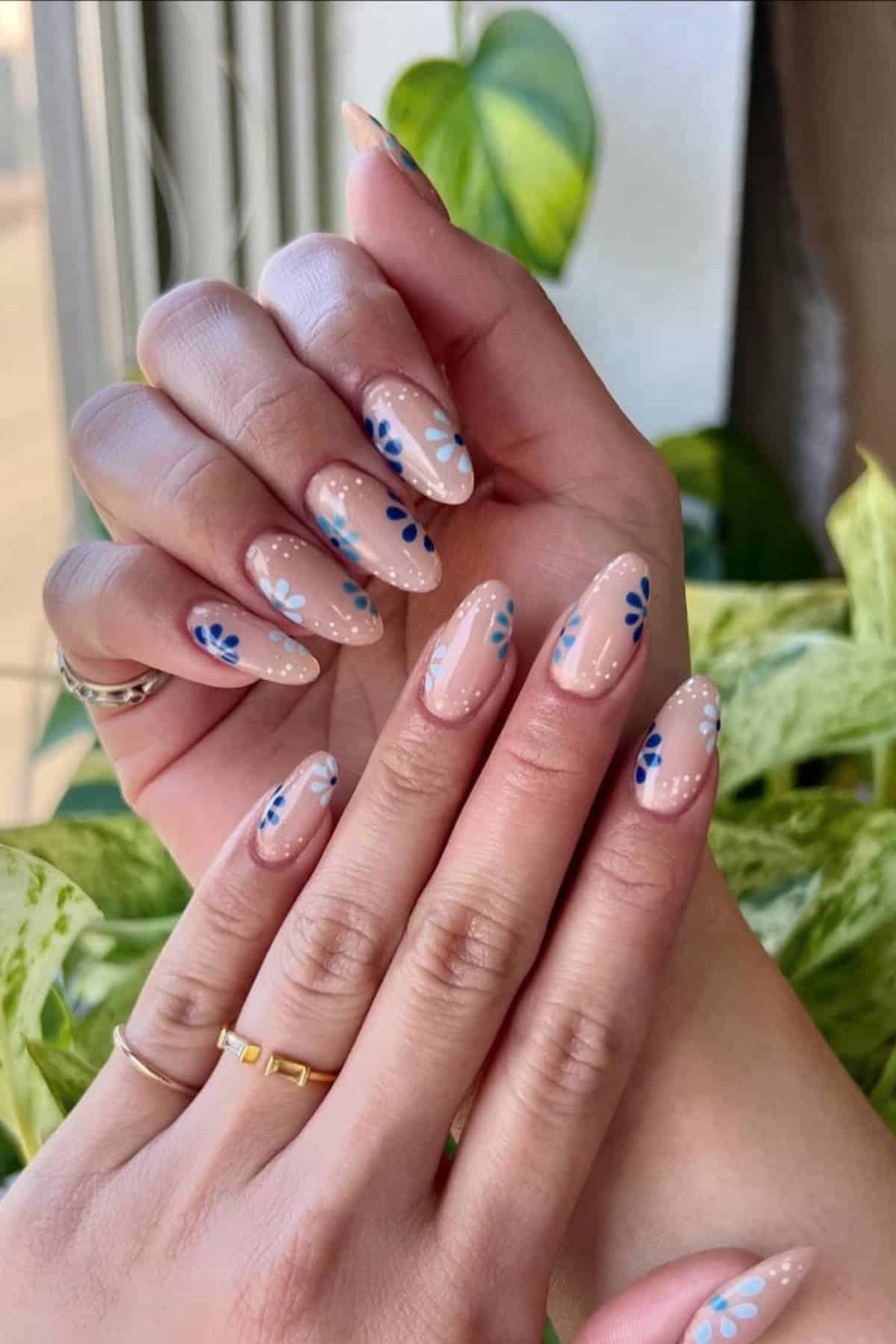 Hands with almond-shaped nails featuring nude polish and blue and white floral nail art, shown against a background of green plants.