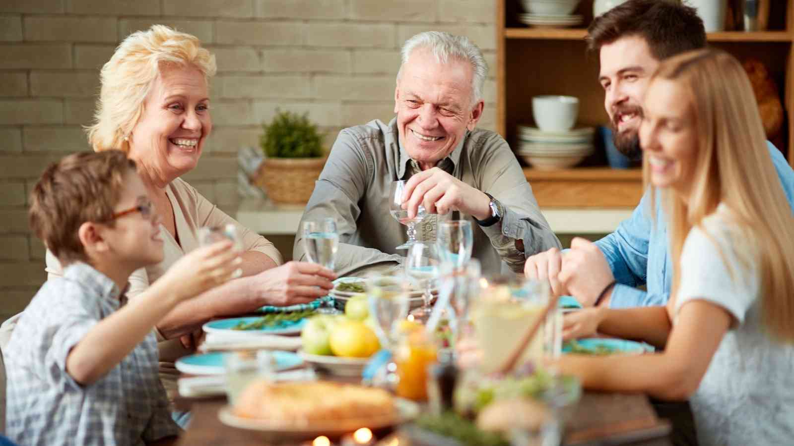Six people of various ages sit around a dining table, smiling and talking while sharing a meal in a home setting.