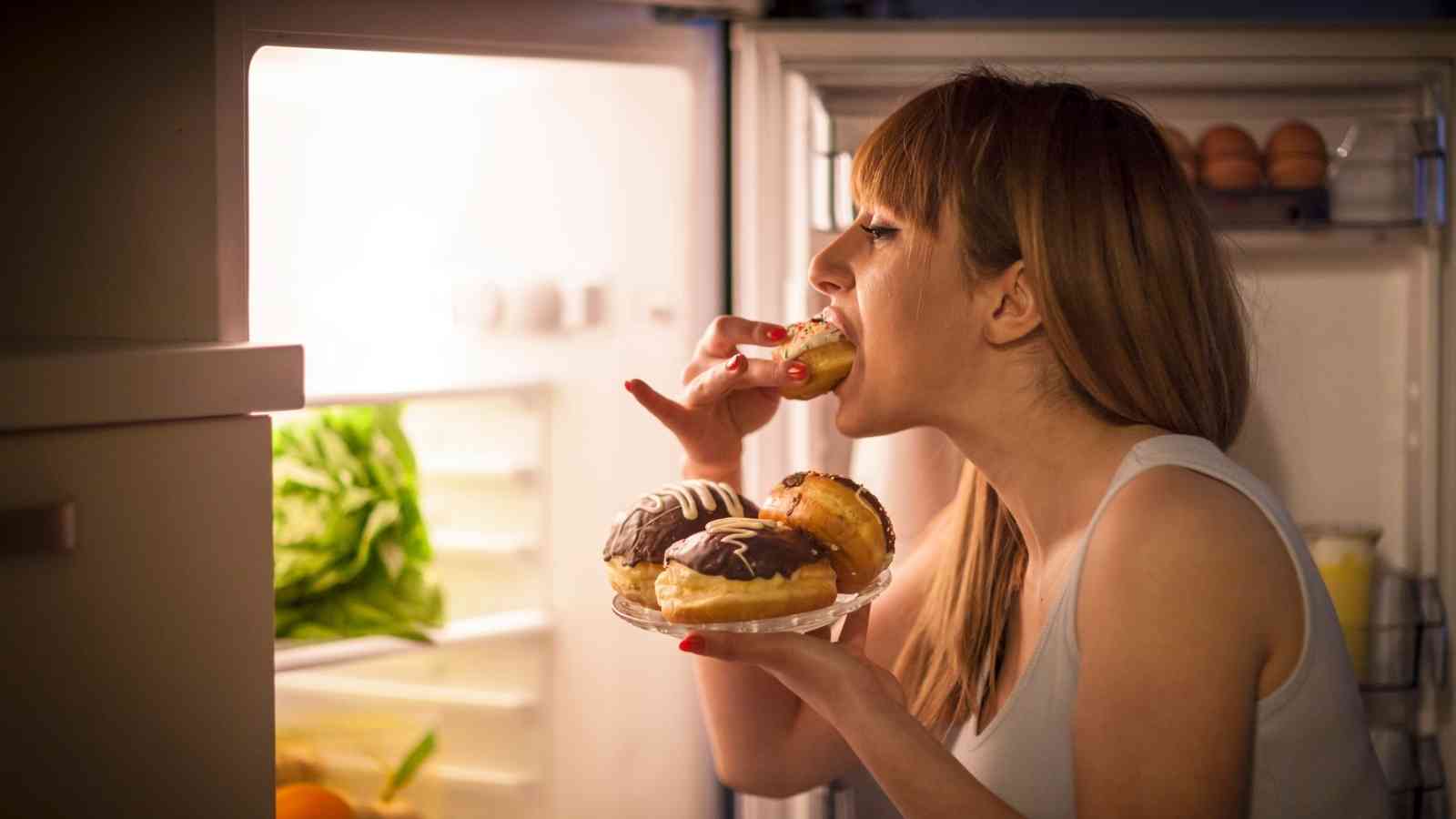 A woman stands in front of an open refrigerator at night, eating donuts and holding a plate with more donuts in her hand.