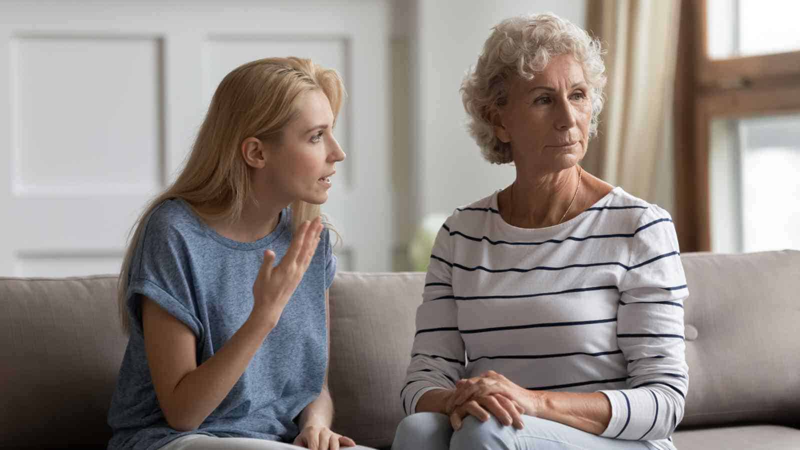 A younger woman appears to be talking seriously to an older woman, who is looking away with a neutral expression. Both are sitting on a couch indoors.