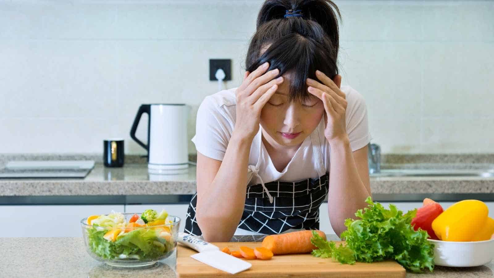 A woman in an apron stands in a kitchen with her head in her hands, surrounded by vegetables and a salad on the counter.