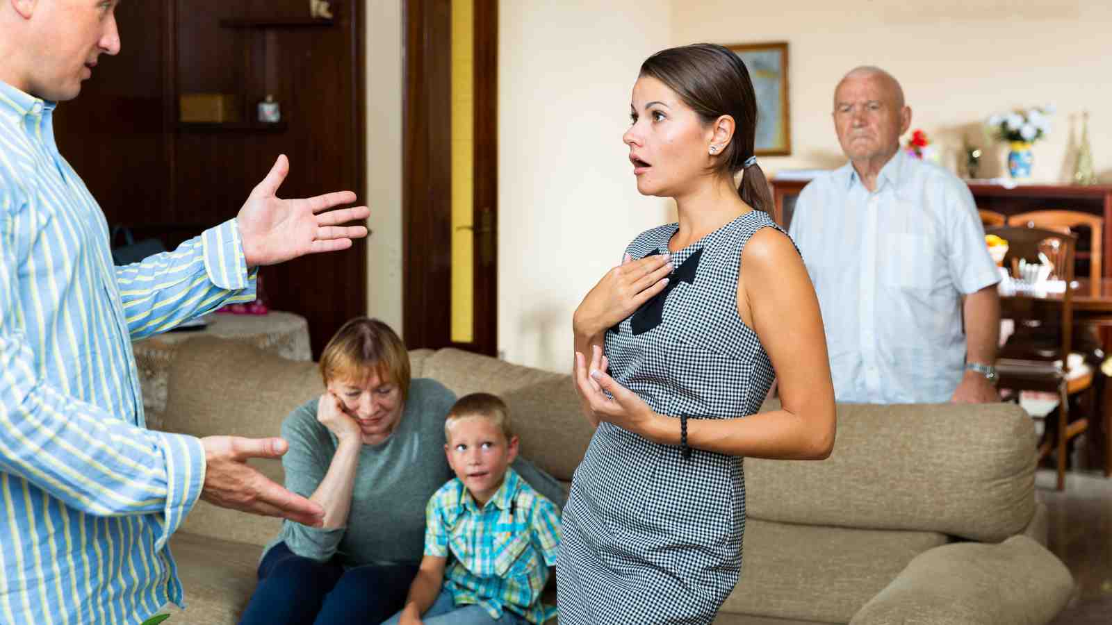 A man and a woman are having a heated discussion in a living room while an older couple and a young boy watch from a couch in the background.