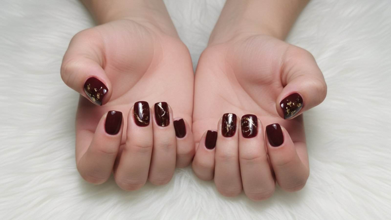 Two hands with short, square-shaped nails painted in dark maroon polish with gold foil accents, resting on a white, fluffy surface.