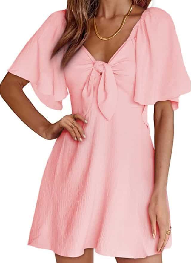 Woman wearing a light pink short dress with flutter sleeves and a front knot detail, accessorized with a gold necklace.