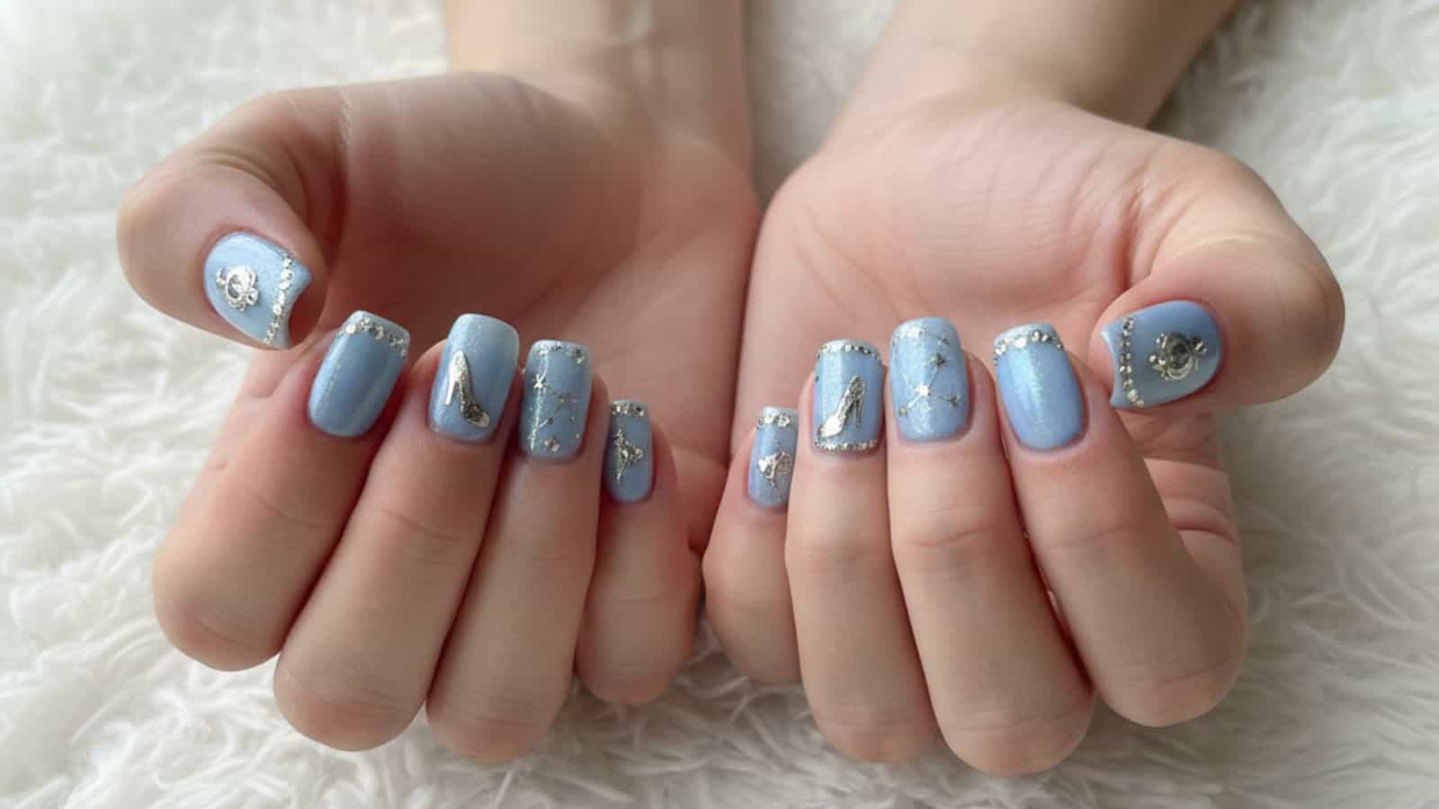 Two hands display long, square-shaped nails painted Cinderella-inspired designs, and silver embellishments, against a white textured background.