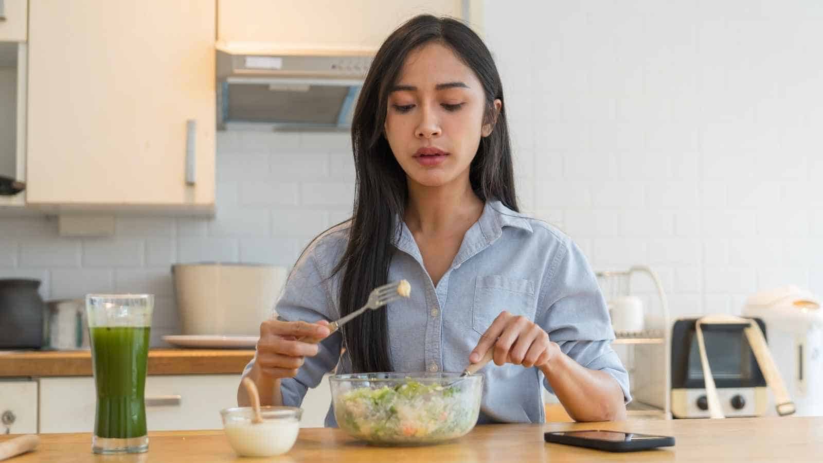 A woman sits at a kitchen table eating salad from a glass bowl, with a glass of green juice, a small bowl of dressing, and a smartphone nearby.