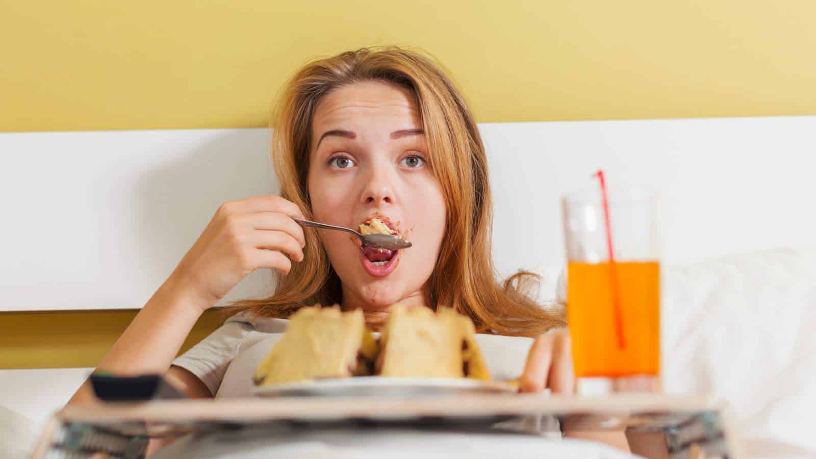 A person sits up in bed eating food from a tray and drinking an orange beverage, with a neutral expression.