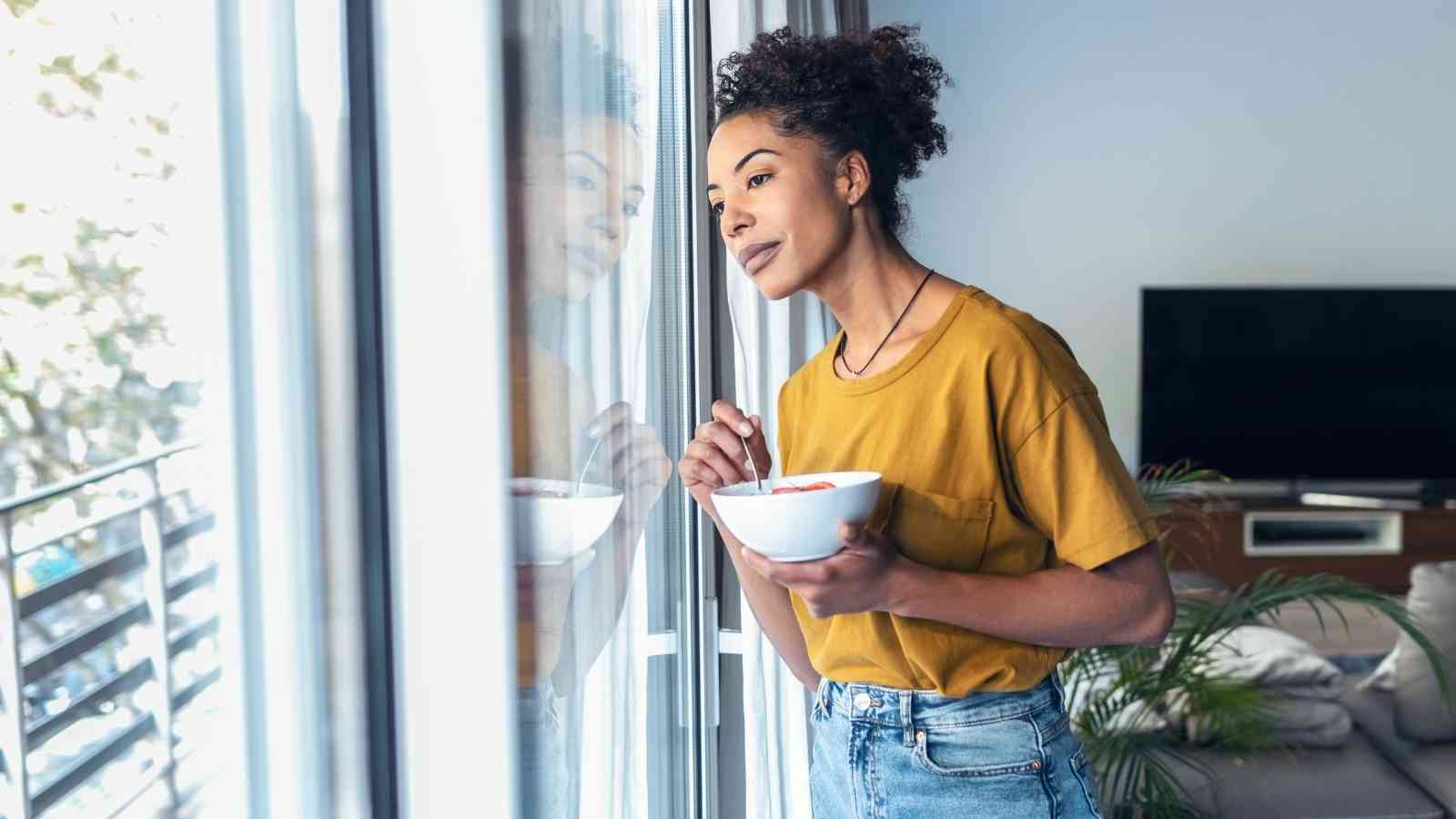 A woman in a yellow shirt stands by a window holding a bowl of food, looking outside in a modern living room.