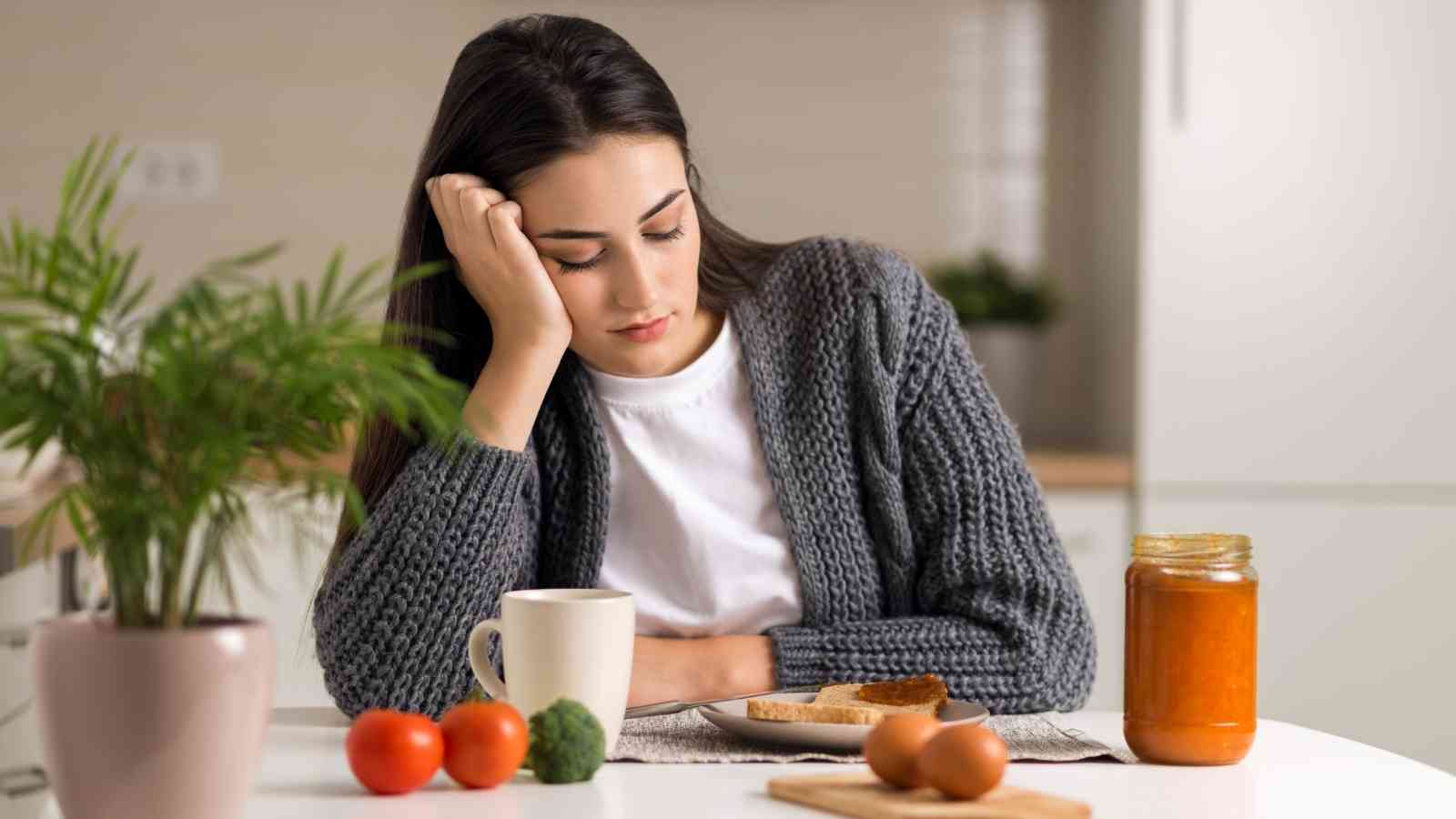 A woman sits at a table with her head resting on her hand, looking at a plate with toast and a jar of orange spread, surrounded by tomatoes, broccoli, and a cup.