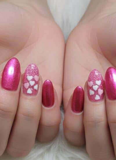 Hands displaying pink, almond-shaped nails with two accent nails on each hand featuring a white heart pattern on a glittery background.
