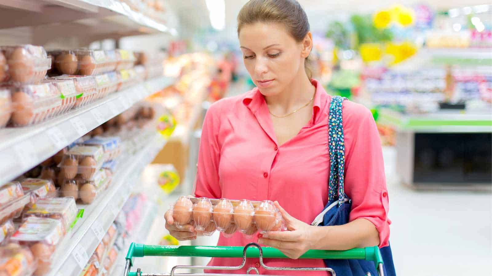 A woman in a pink shirt examines a carton of eggs in a grocery store aisle, holding a shopping cart with groceries.