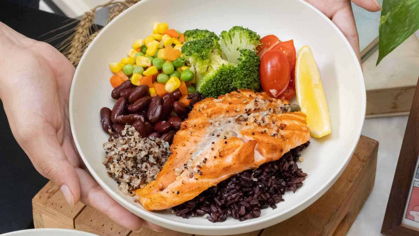 A bowl with grilled salmon, black rice, quinoa, kidney beans, mixed vegetables, broccoli, grape tomatoes, and a lemon wedge.