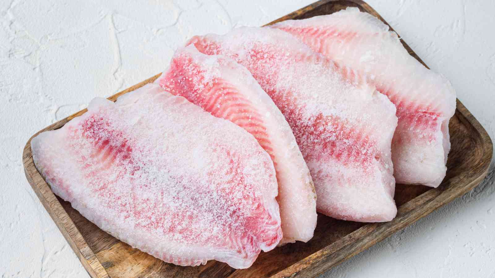 Four frozen fish fillets with visible ice crystals are arranged on a wooden tray against a light textured background.