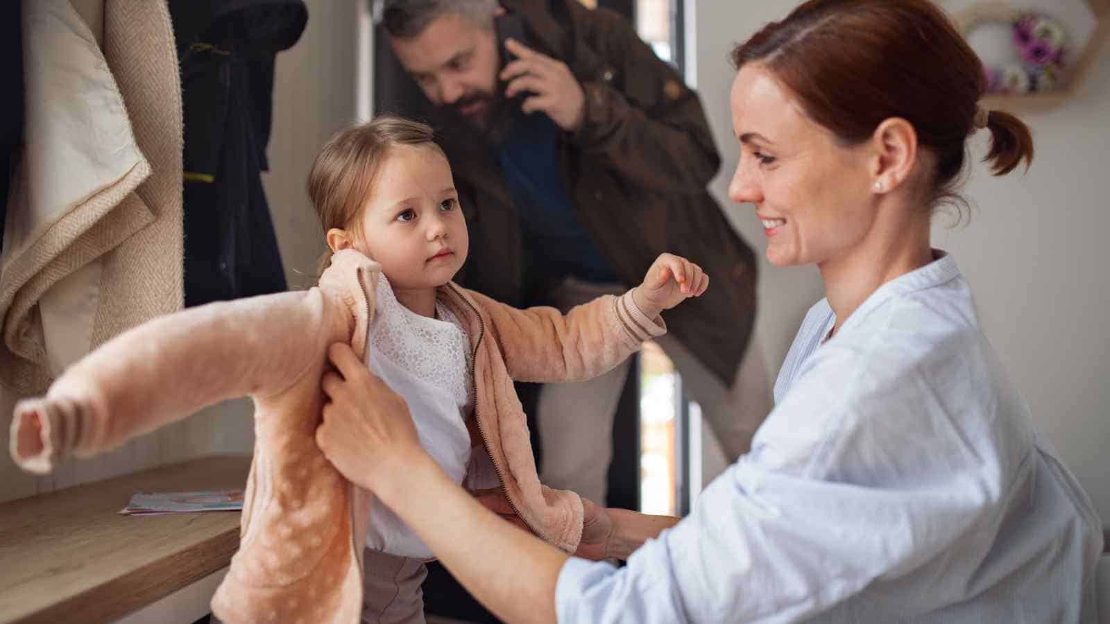 A woman helps a young girl put on a jacket while a man in the background speaks on the phone.