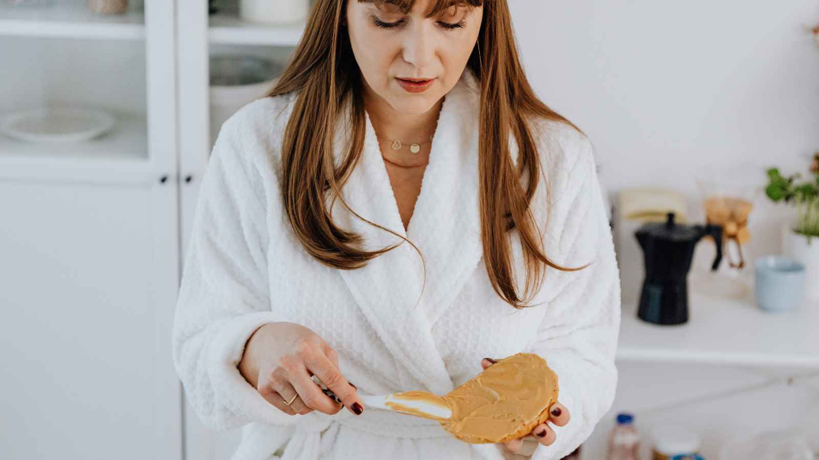 Woman in a white robe spreads peanut butter on bread in a kitchen.