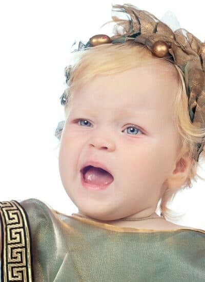 A young child wearing a gold laurel wreath and toga gestures with an open mouth, as if speaking or shouting, against a white background.