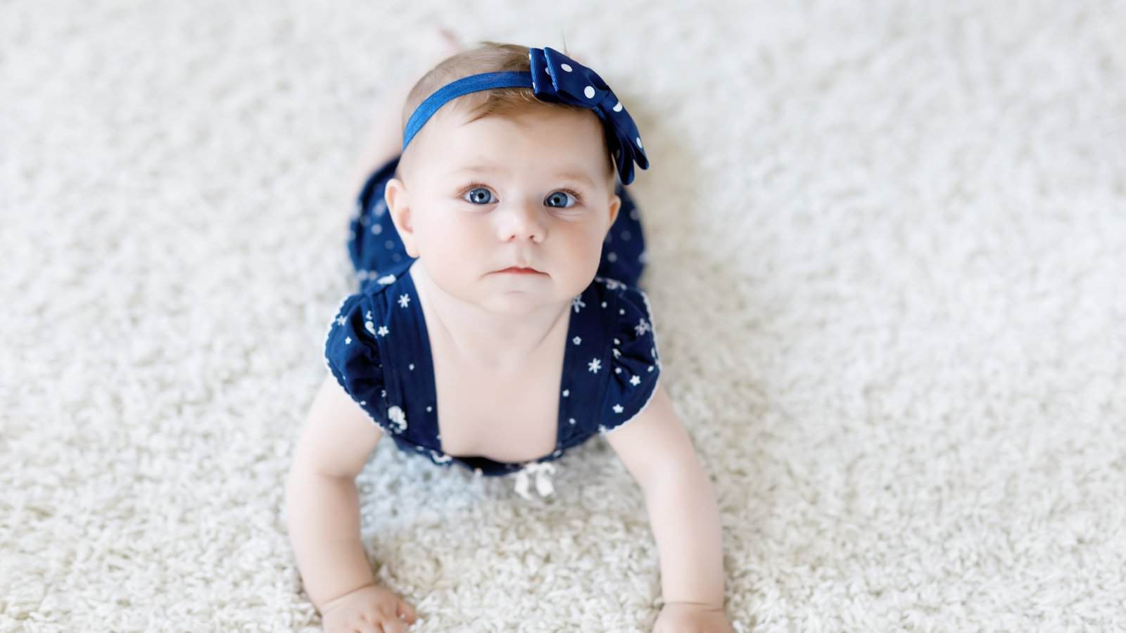 A baby wearing a blue headband with white polka dots and a matching blue outfit lies on a white carpet, looking up.