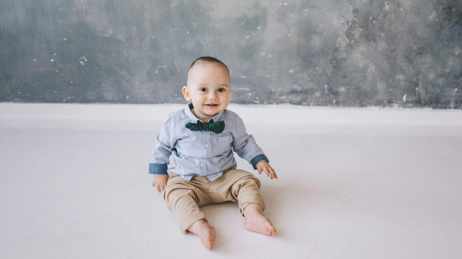 A baby wearing a blue shirt, green bow tie, and beige pants sits barefoot on a white floor against a gray textured wall.
