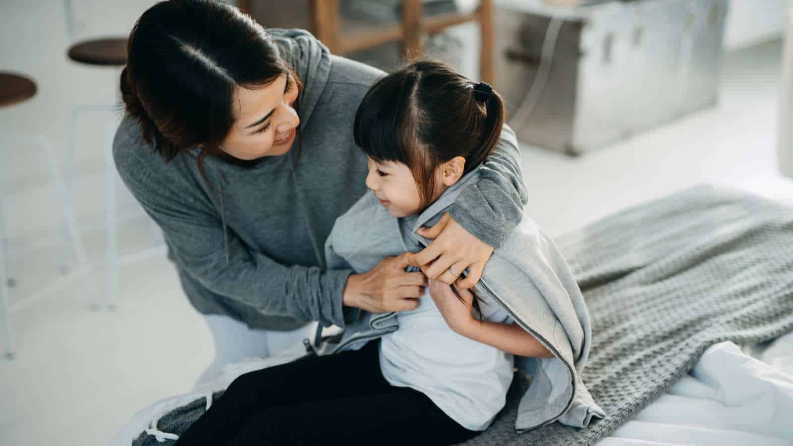 An adult helps a young child put on a gray sweater while sitting on a bed in a bright, modern room.