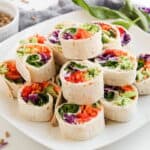 A white plate with neatly stacked veggie pinwheel wraps filled with colorful vegetables, next to a small bowl of seeds and scattered spinach leaves.