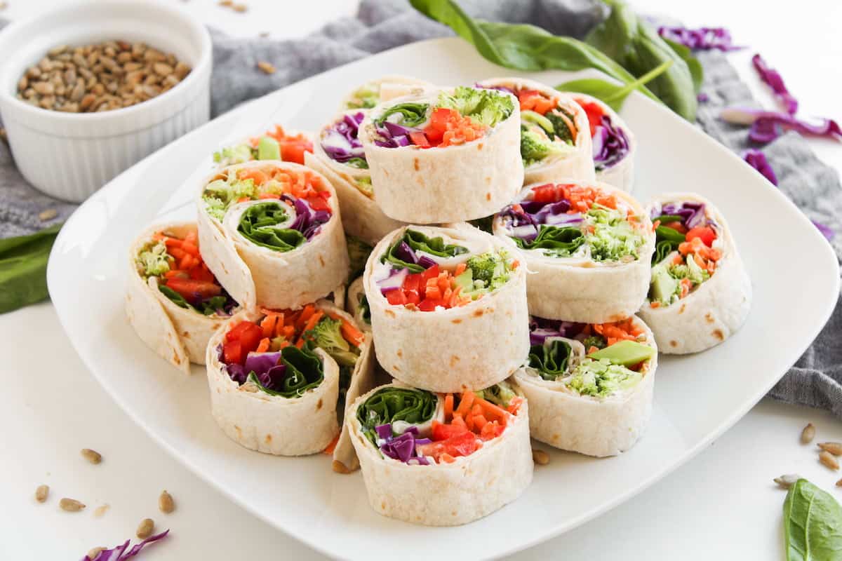 A white plate with neatly stacked veggie pinwheel wraps filled with colorful vegetables, next to a small bowl of seeds and scattered spinach leaves.