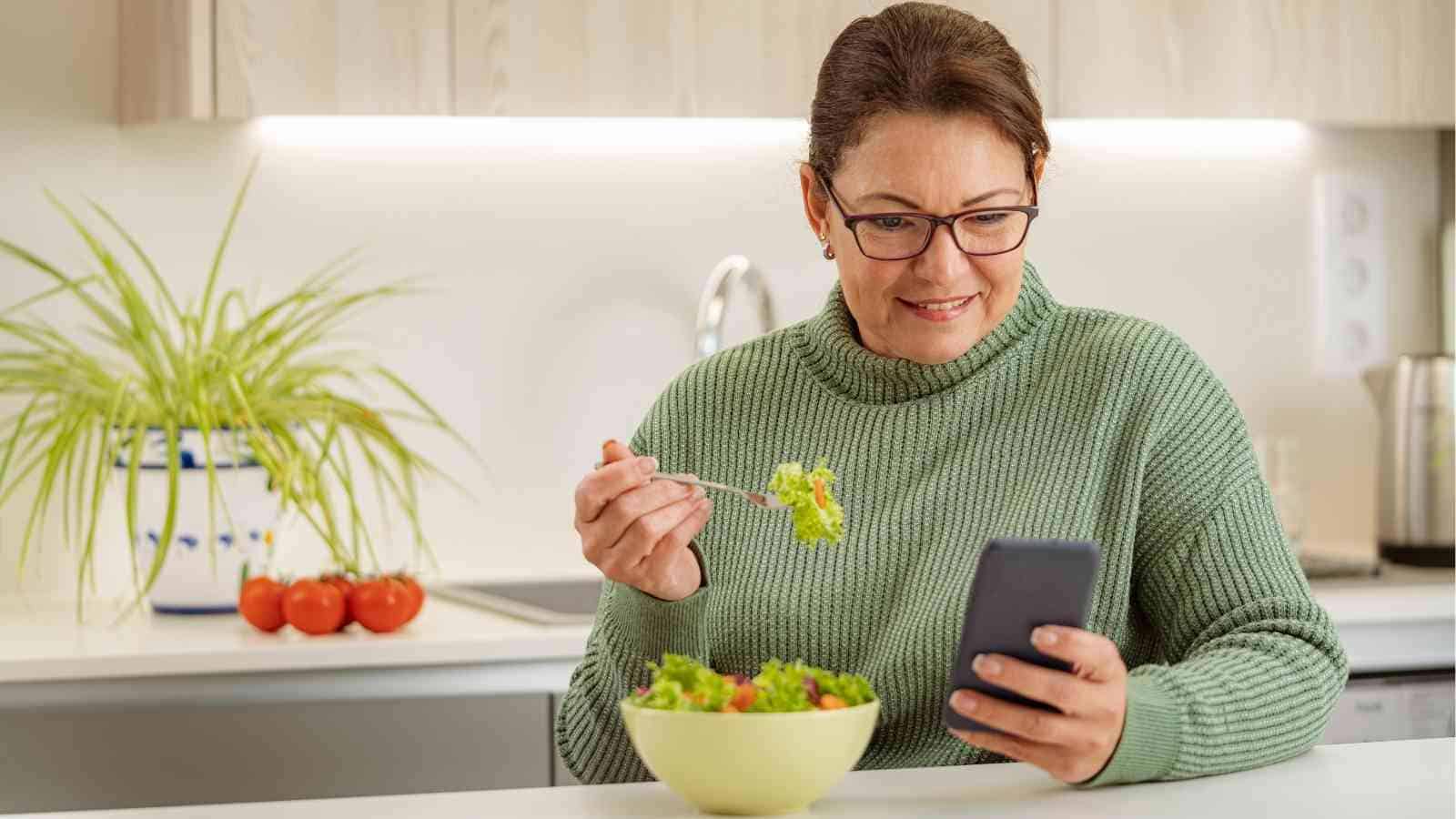 Woman wearing glasses and a green sweater sits at a kitchen counter, eating salad from a bowl and looking at her smartphone.
