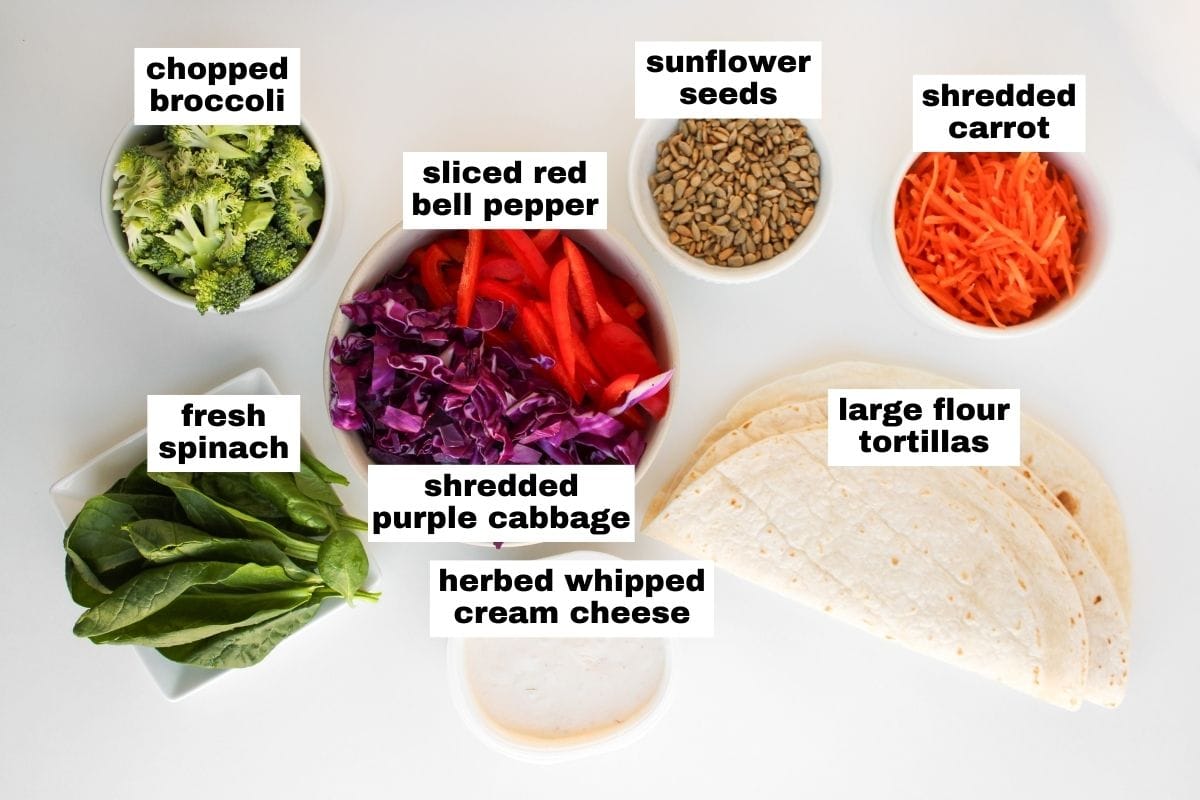 A flat lay of fresh spinach, chopped broccoli, shredded purple cabbage, sliced red bell pepper, shredded carrot, sunflower seeds, herbed whipped cream cheese, and large flour tortillas.