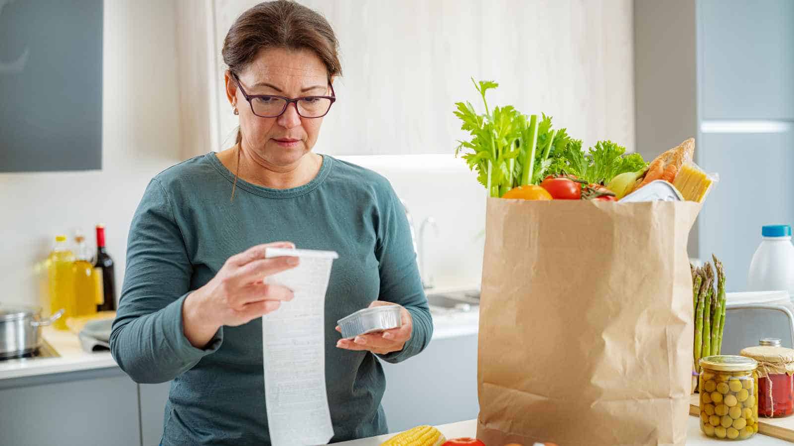 A woman stands in a kitchen holding a receipt and a credit card next to a paper bag filled with groceries.
