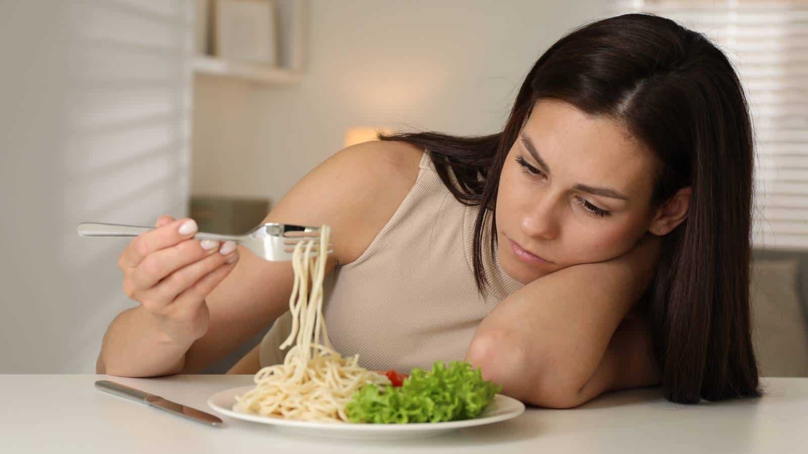 A woman sits at a table, resting her head on her arm and looking unenthusiastically at a forkful of spaghetti with salad on a plate in front of her.