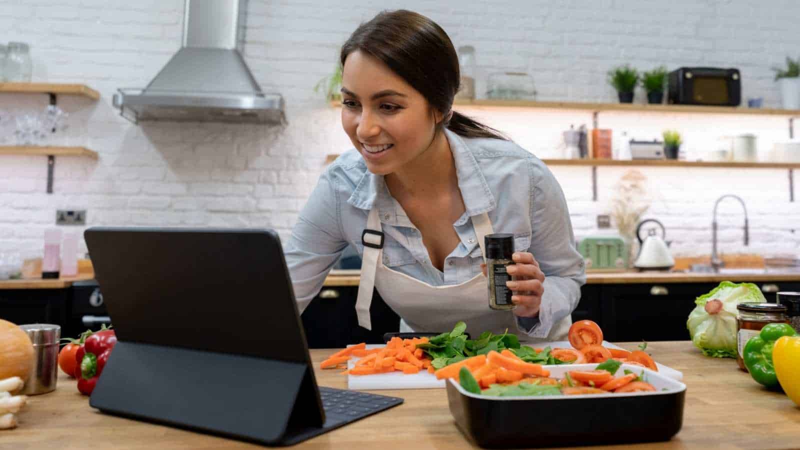 A woman in a kitchen chops vegetables and looks at a tablet, holding a spice jar, with kitchen utensils and ingredients around her.
