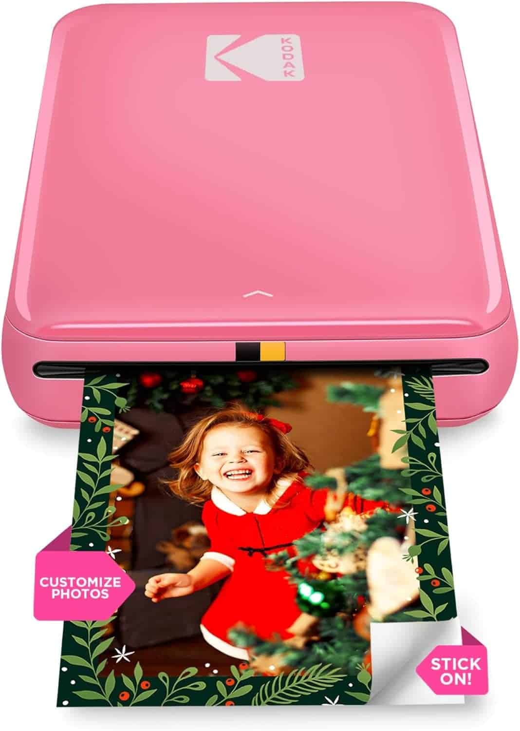 A pink Kodak photo printer prints a holiday-themed photo of a smiling child in a red dress next to a Christmas tree. Labels read "Customize Photos" and "Stick On!.