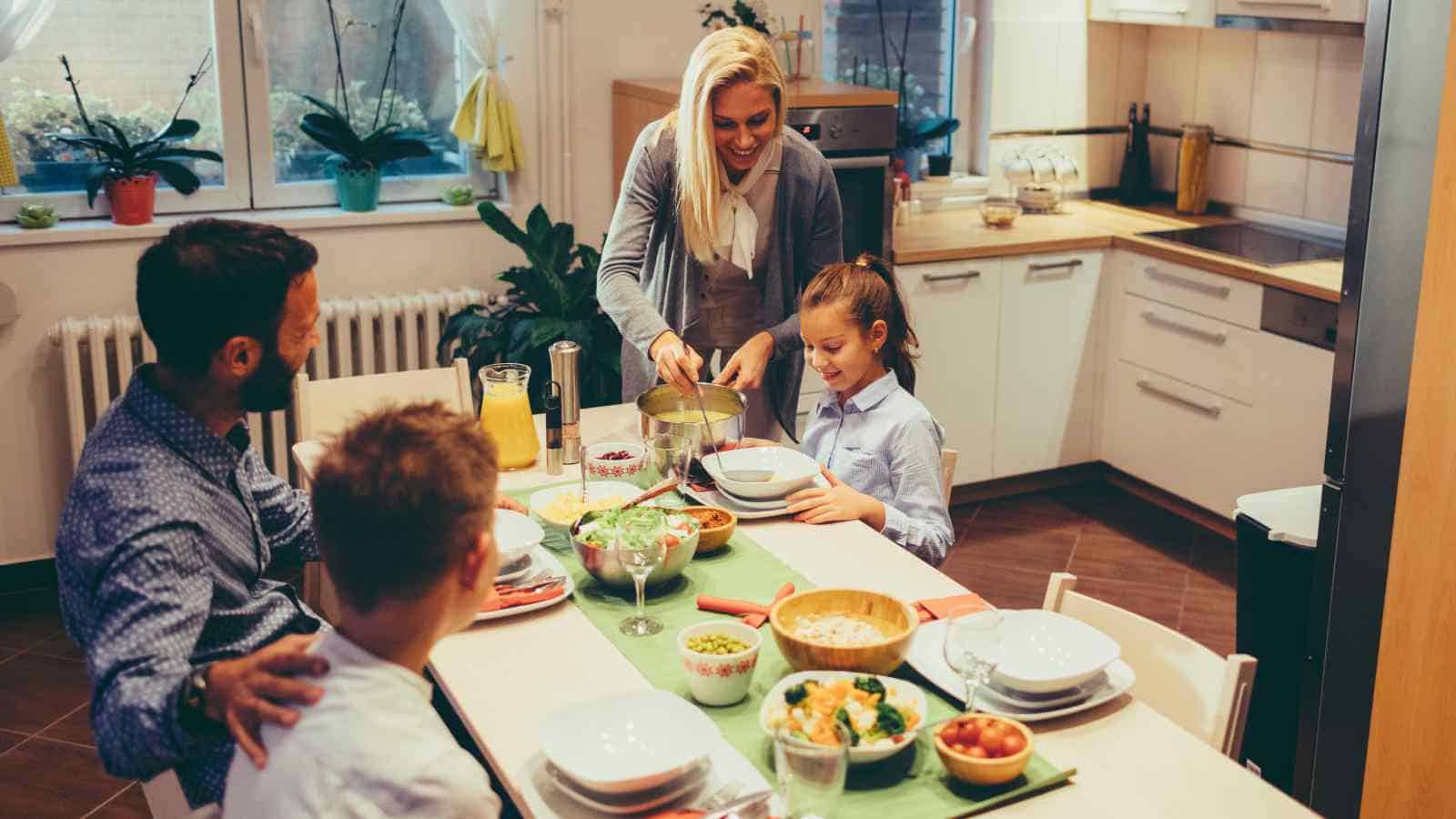 A family of four sits at a kitchen table set for a meal while a woman serves food to a young girl; various dishes and drinks are arranged on the table.