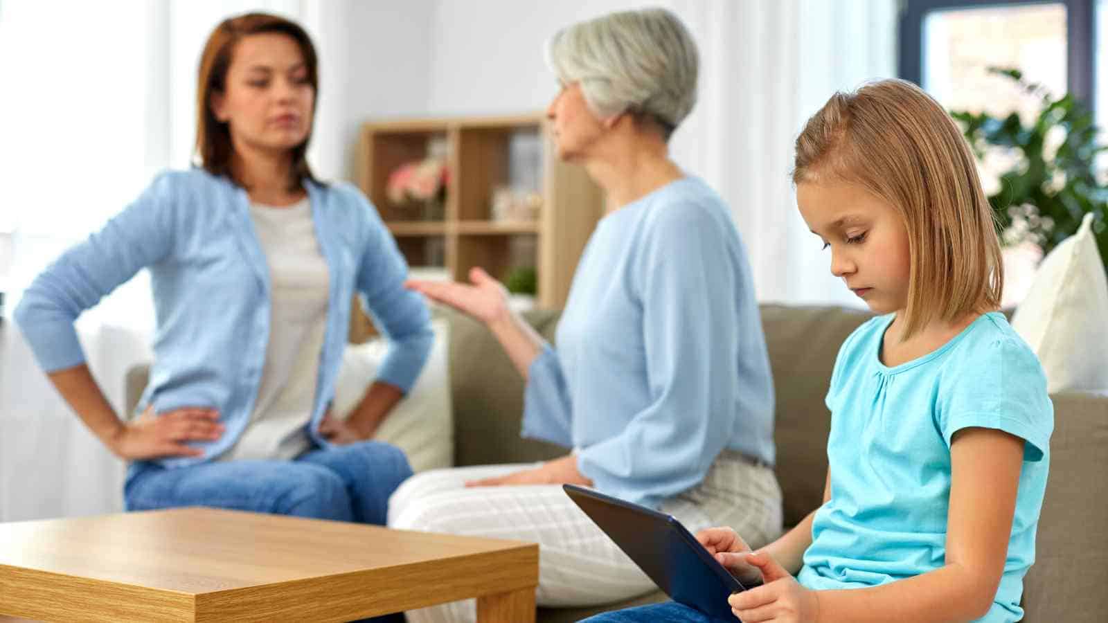 A girl sits on a couch using a tablet while two women in the background appear to be having a serious conversation.