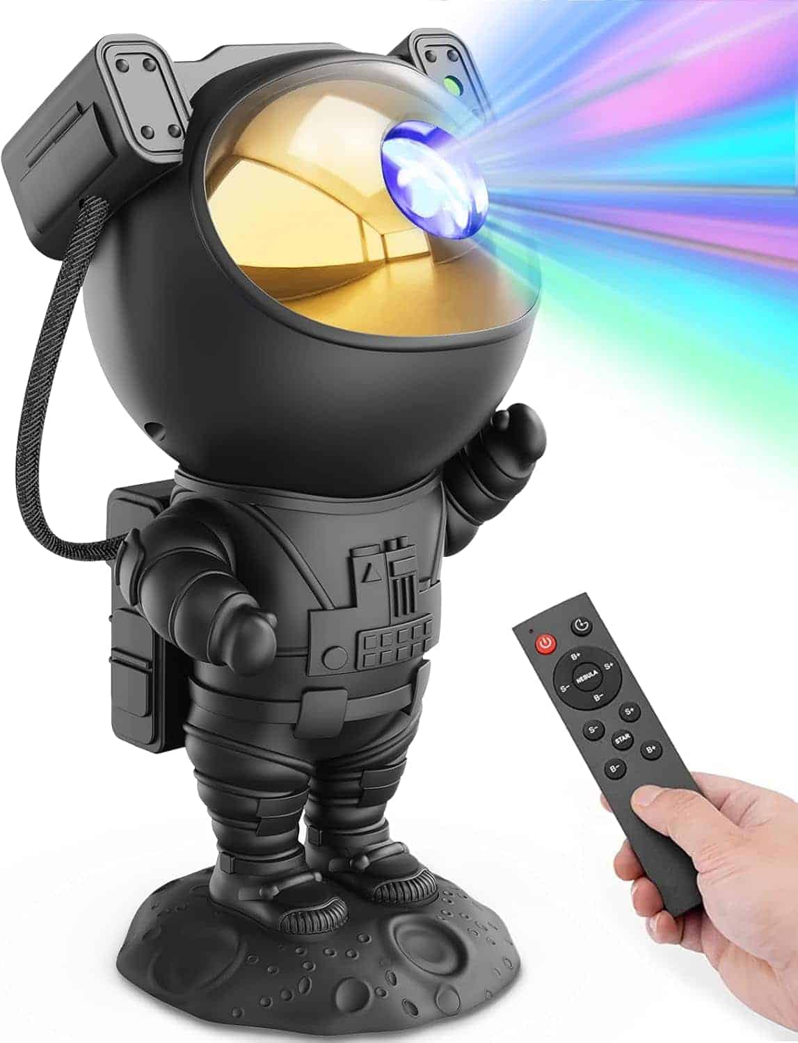 A hand holds a remote control, aiming it at a black astronaut-shaped projector lamp that emits colorful light beams from its helmet visor.