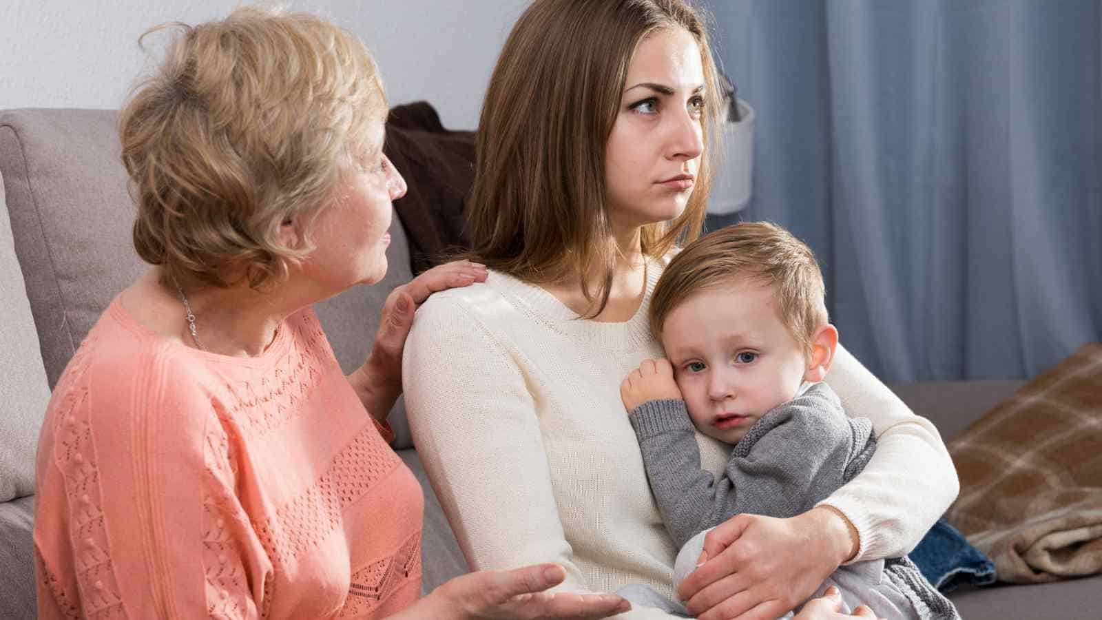 Three people sit on a couch; an older woman is talking, a young woman looks serious while holding a small child who appears worried.