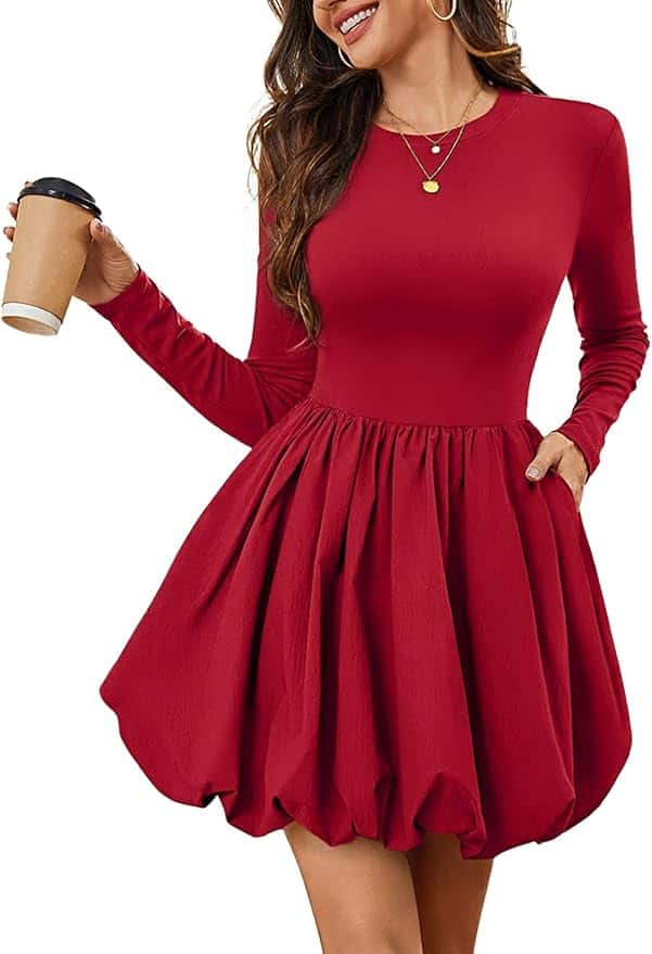 Woman wearing a long-sleeve, red, knee-length dress with a fitted bodice and flared skirt, holding a coffee cup in one hand and the other hand in her pocket.
