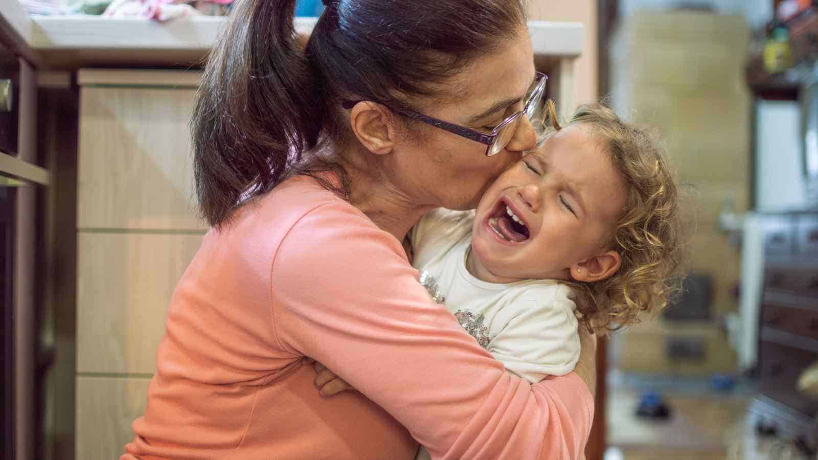 A woman wearing glasses hugs and kisses a crying toddler indoors, comforting the child who appears upset.