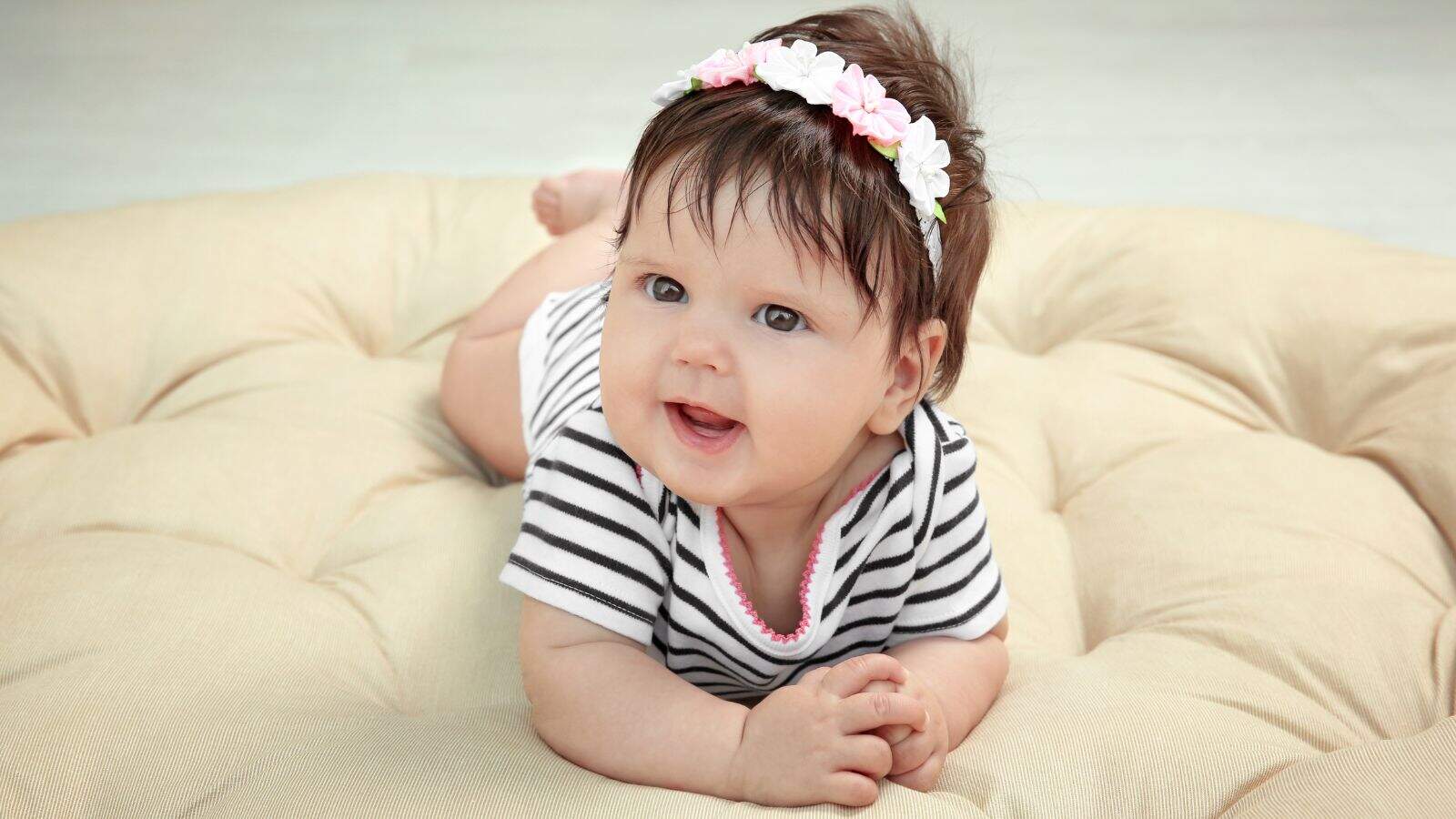 A baby wearing a striped onesie and a floral headband lies on their stomach on a beige cushion, looking up and smiling.