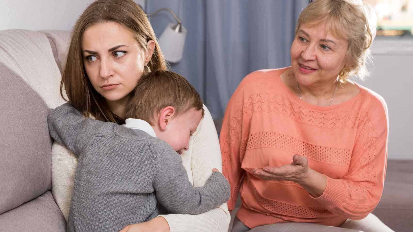 A woman sits on a couch holding a crying child while an older woman beside them gestures, appearing to speak or explain something.