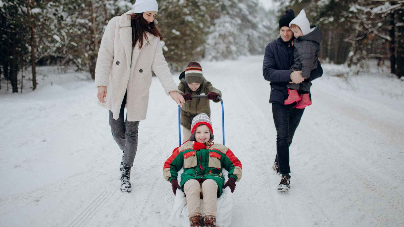 A family walks in the snow; a child sits on a sled, another child pushes, and two adults walk alongside, one carrying a small child.