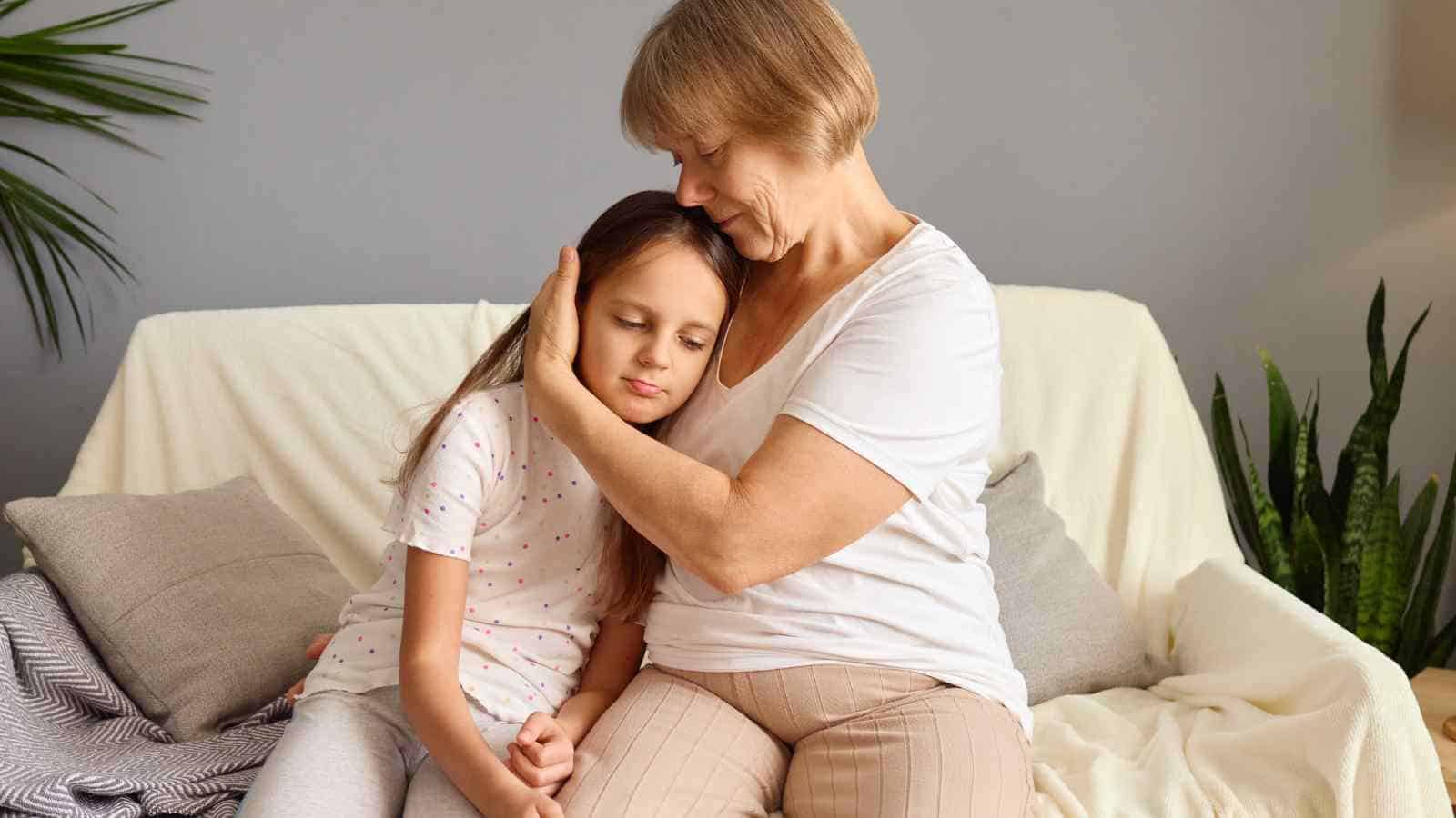 An older woman sits on a couch, gently hugging a young girl who leans against her with closed eyes. Both appear calm and relaxed in a cozy, home setting with plants in the background.