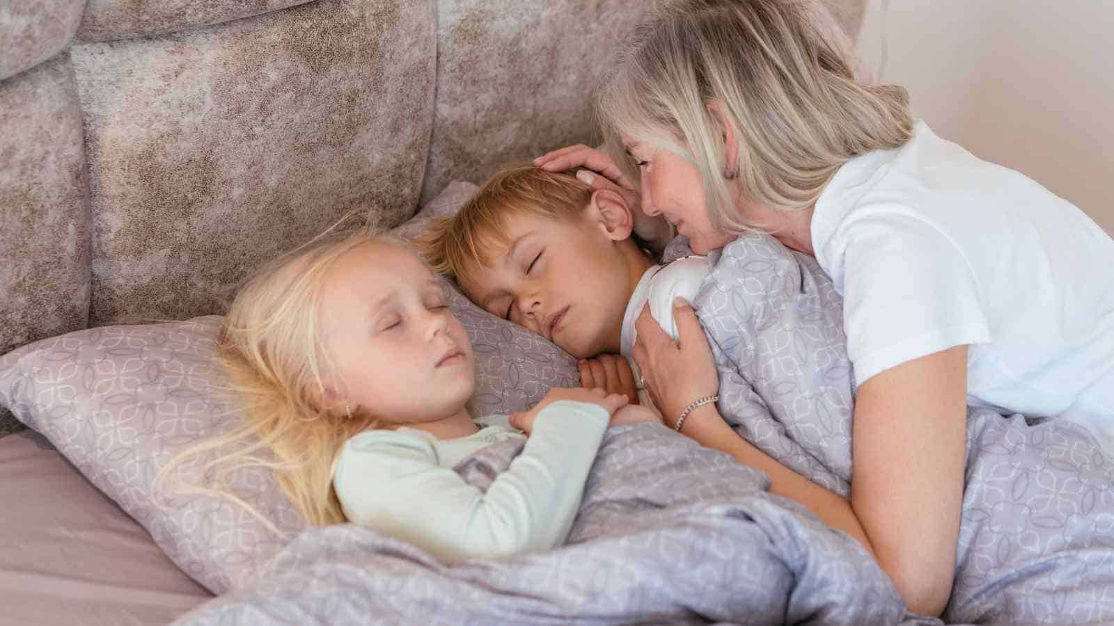 An older woman gently tucks two young children into bed as they sleep, all covered by a grey patterned blanket.