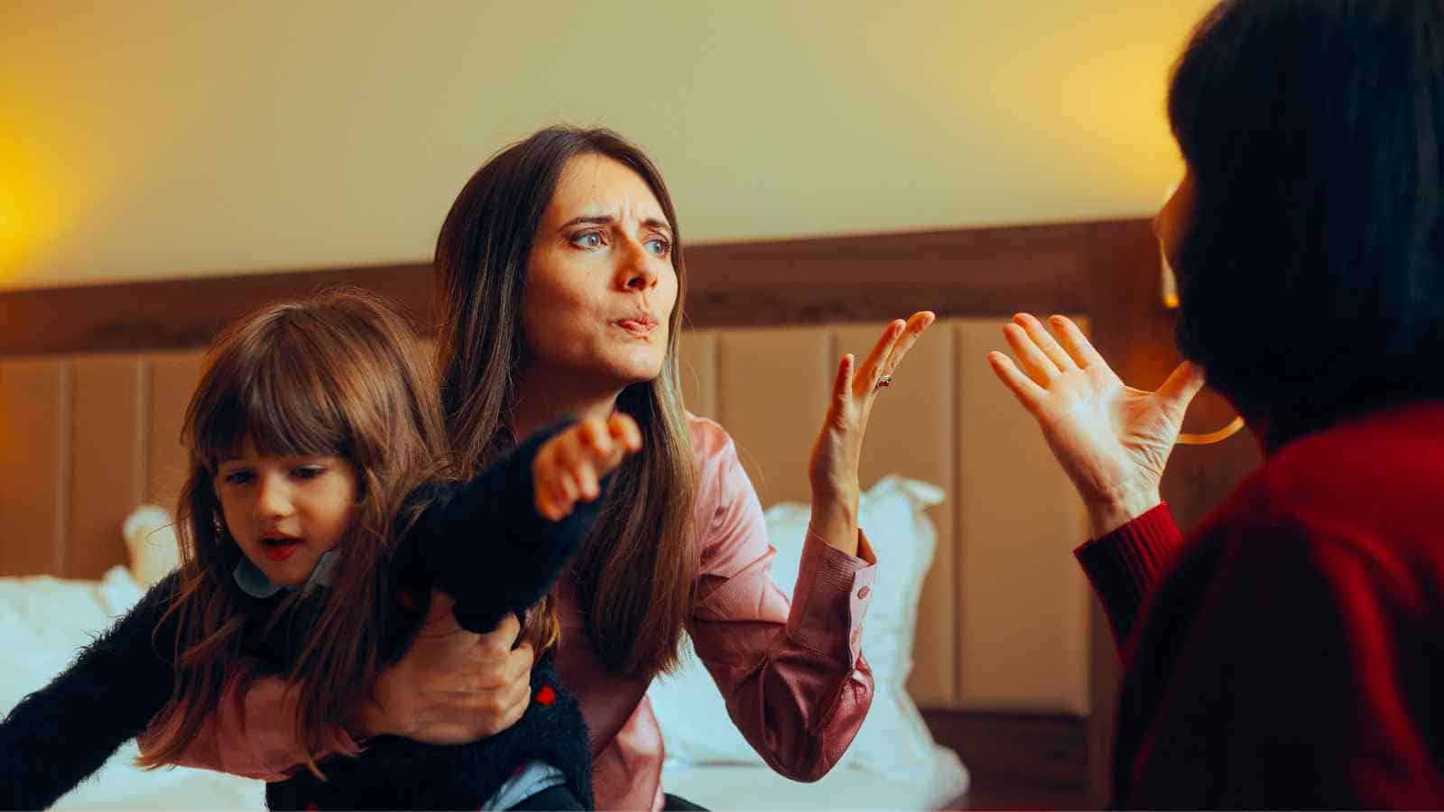 A woman holding a young child gestures emotionally toward another woman in a heated conversation in a bedroom.