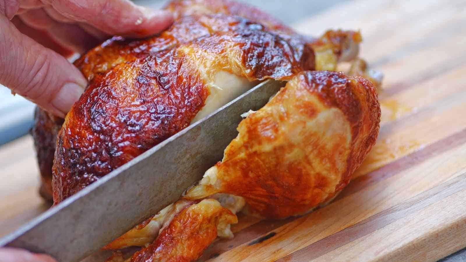 A person slices a roasted chicken with a large knife on a wooden cutting board.