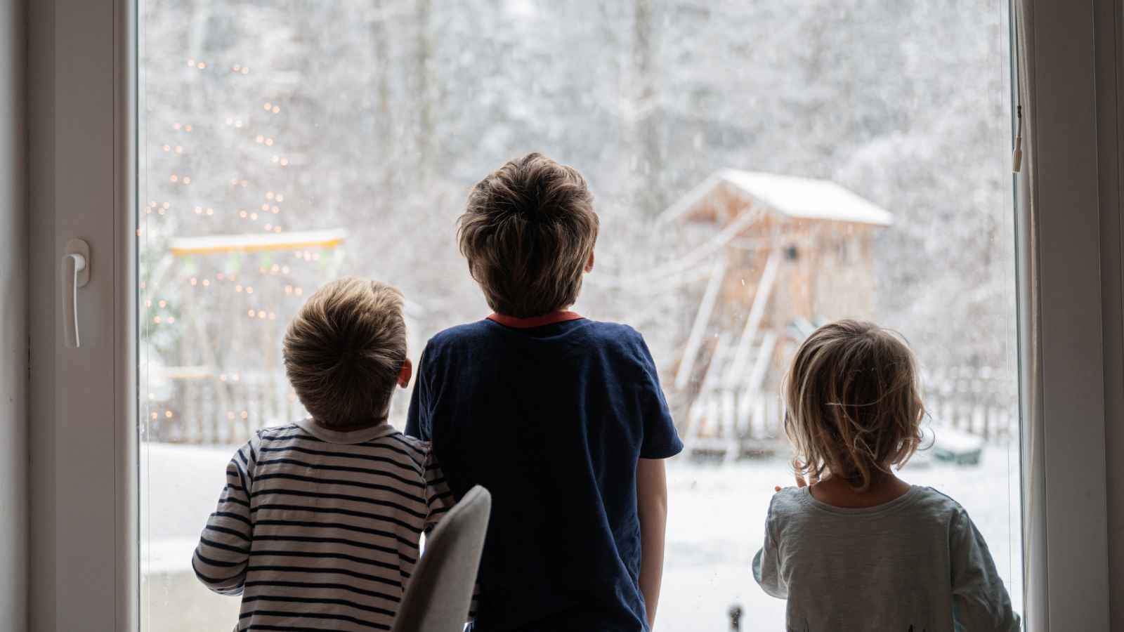 Three children stand indoors looking out a window at a snowy backyard with a wooden playhouse and trees covered in snow.