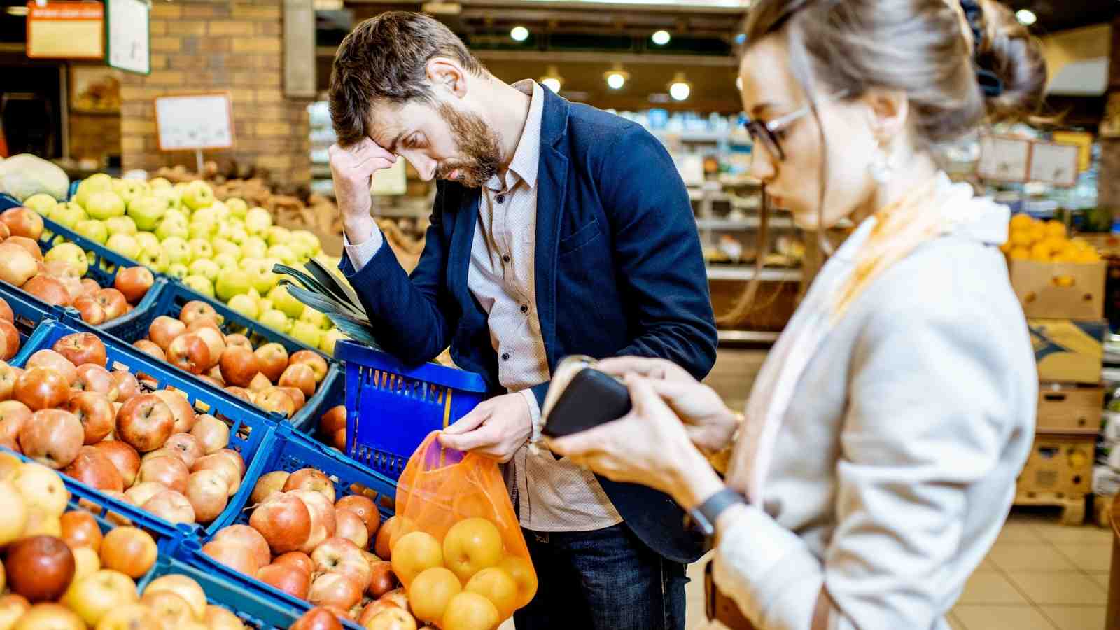 A man looks stressed while selecting apples at a grocery store, holding a basket, as a woman beside him checks a wallet.