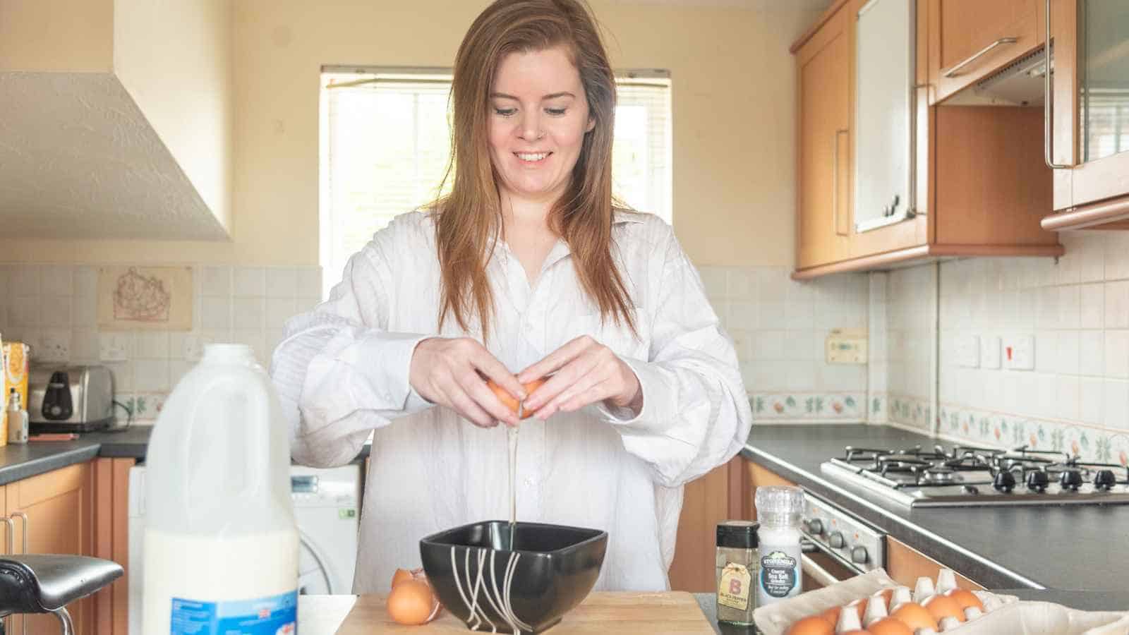 A person cracks an egg into a black bowl in a kitchen, with eggs, milk, and seasonings on the counter.